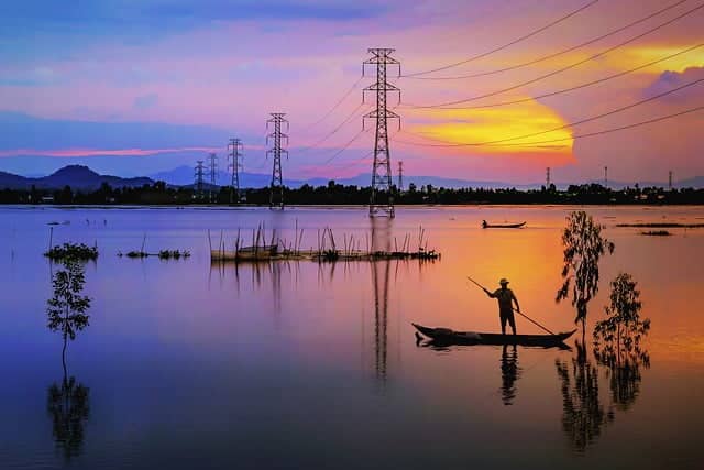 Page A propos un coucher de soleil réverbère sur une étendue d'eau où un homme se tient debout sur une barque qu'il propulse à laide d'une perche. On voit sur le fond des pilonnes électriques qui réverbèrent leurs ombres sur l'eau dans la page A propos