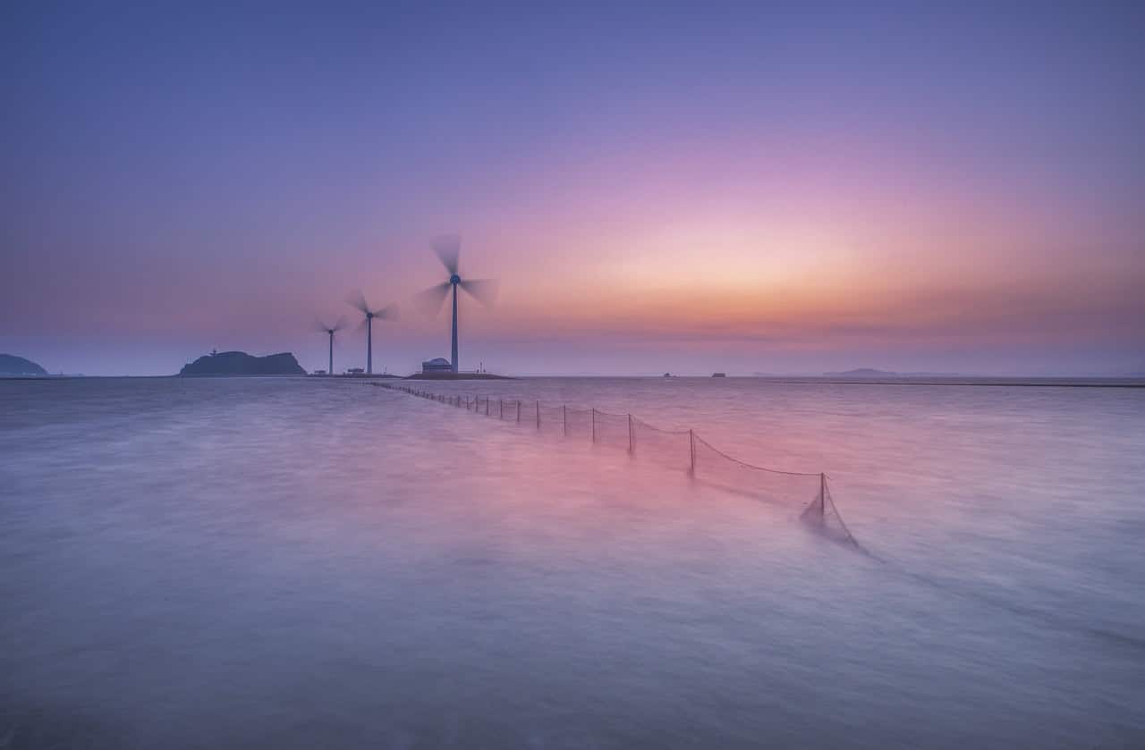 page Projets photo montrant un projet realisable avec les énergies renouvelables .c'est un paysage marin avec un soleil caché produisant une Lumiere violette qui reflète dans l'eau dans laquelle une barrière très longue apparait.au loin sur une ile trois éoliennes semblent tourner à grande vitesse.